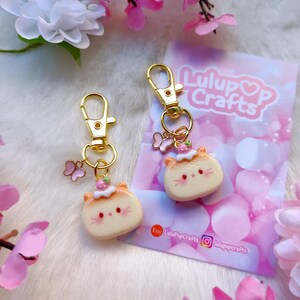 May include: Two gold keychains with a cute cartoon cat charm. Each charm has a pink flower crown, pink cheeks, and a white face. The charms are attached to gold key rings with a small butterfly charm.