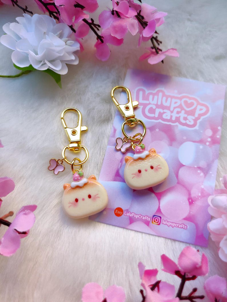 May include: Two gold keychains with a cute cartoon cat charm. Each charm has a pink flower crown, pink cheeks, and a white face. The charms are attached to gold key rings with a small butterfly charm.