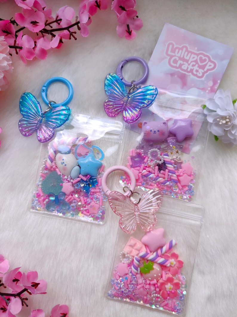 May include: Several clear plastic bags filled with colorful beads and charms, including stars, hearts, and flowers. Each bag has a butterfly-shaped keychain attached. The bags are in shades of pink, blue, and purple, with the text "Lulup Crafts" on a pink card.