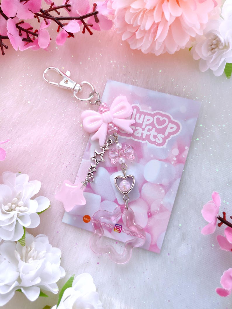 A kawaii pastel keychain featuring a soft pink bow charm with pearl accents, dangling pastel star and flower charms, silver star links, and a heart-shaped frame with a pearl inside. Displayed on a dreamy pink and white background with delicate faux flowers for a cute and whimsical aesthetic