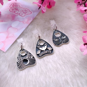 May include: Three silver charm pendants with black enamel designs. The charms are shaped like a witch's board with the words "Yes", "No", and "Goodbye" on them. One charm has a bat and spiderwebs, another has a crescent moon, and the third has a black cat face.