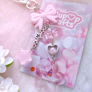 May include: A pink keychain with a silver clasp, featuring a pink bow, star charms, and a flower-shaped charm. The keychain is attached to a card with the text "lupop crafts" and social media handles.