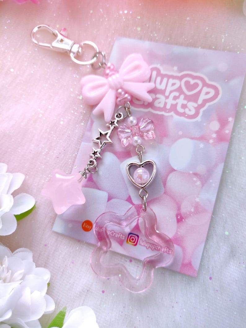 May include: A pink keychain with a silver clasp, featuring a pink bow, star charms, and a flower-shaped charm. The keychain is attached to a card with the text "lupop crafts" and social media handles.