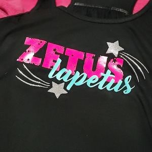 Puede incluir: Camiseta de tirantes negra con las palabras "ZETUS lapetus" en letras rosa y turquesa con estrellas de purpurina plateada y estrellas fugaces.