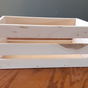 Pode incluir: Caixa de madeira retangular pintada de branco, com grão e nós de madeira visíveis. A caixa tem um design de ripas nas laterais e é construída com pregos visíveis. O interior é de madeira sem pintura. Adequada para armazenamento ou exibição.