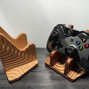May include: Two wooden stands for video game controllers. One stand is a simple design with two slots, the other is more elaborate with a wavy design. A black and grey camouflage Xbox controller is on the stand.
