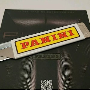 May include: A white utility knife with a silver blade and a yellow label that says "PANINI" in red letters. The knife is lying on top of a black and white patterned surface with the word "PANINI" printed in black.