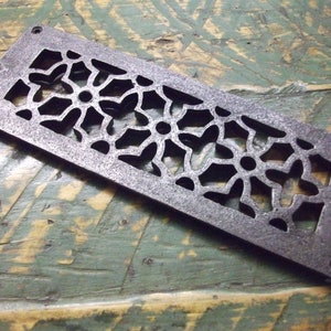 May include: A rectangular cast iron vent cover with a decorative floral pattern. The vent cover is a dark gray color and has a slightly textured surface.