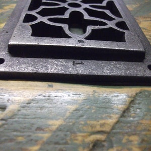 May include: A silver metal vent cover with a decorative, geometric design. The vent cover has a rectangular shape and is slightly raised from the surface. The vent cover has a small, rectangular opening in the center.