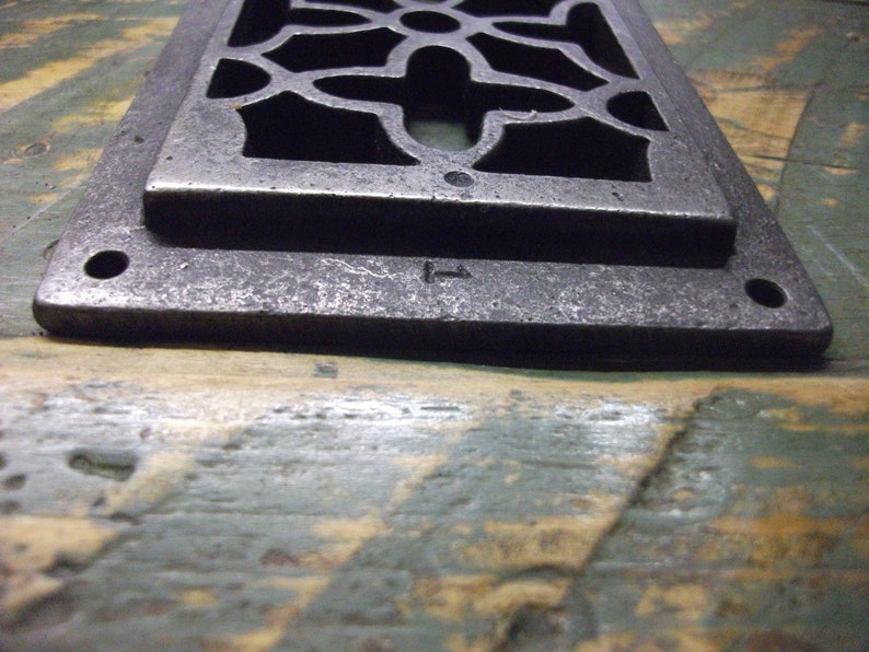 May include: A silver metal vent cover with a decorative, geometric design. The vent cover has a rectangular shape and is slightly raised from the surface. The vent cover has a small, rectangular opening in the center.