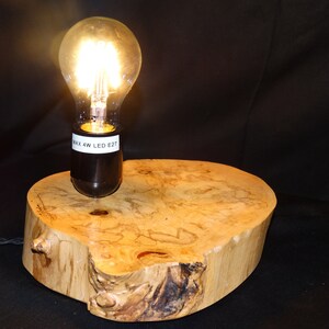 May include: A small wooden table lamp with a light bulb turned on. The lamp is made from a natural wood slice with a black electrical socket and cord. The label on the socket reads "MAX 4W LED E27".