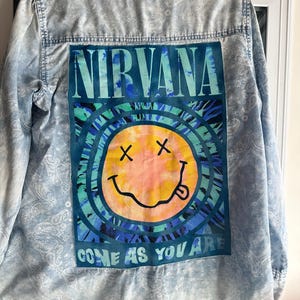 May include: Light blue denim shirt with a Nirvana graphic on the back. The graphic features the band's name in teal, a smiley face with crossed-out eyes and a tongue, and the phrase "COME AS YOU ARE". The shirt has a paisley pattern.