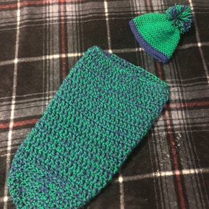 May include: A green and blue crocheted baby sleeping bag and hat set. The sleeping bag is long and narrow, with a textured pattern. The hat has a small pom-pom on top.