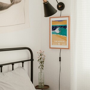 May include: A black and white illustration of a whale tail on a beige tapestry hanging on a white wall. A black wall lamp with a black shade is mounted on the wall. A framed poster with the text "THE WEDGE" and a colorful illustration of a wave is hanging on the wall. A bed with a white and black checkered duvet cover and a white pillow is in the foreground.