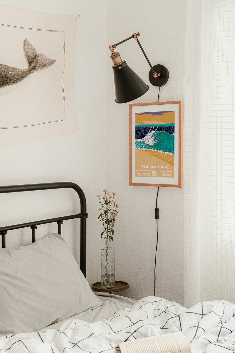 May include: A black and white illustration of a whale tail on a beige tapestry hanging on a white wall. A black wall lamp with a black shade is mounted on the wall. A framed poster with the text "THE WEDGE" and a colorful illustration of a wave is hanging on the wall. A bed with a white and black checkered duvet cover and a white pillow is in the foreground.