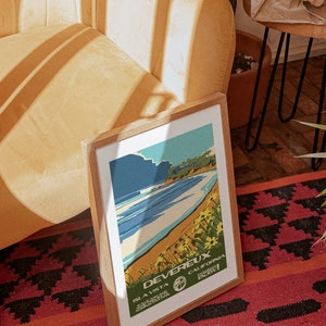 May include: A framed vintage travel poster with a blue and white illustration of a beach scene. The poster reads "Devereux, Isla Vista, California, Enjoy!"