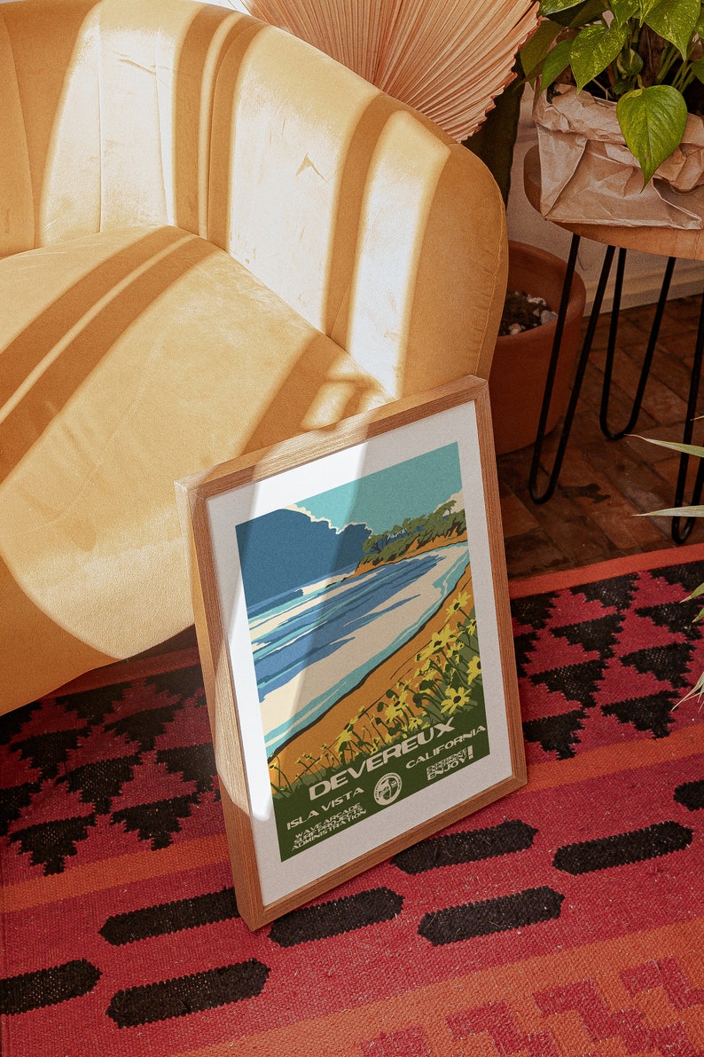 May include: A framed vintage travel poster with a blue and white illustration of a beach scene. The poster reads "Devereux, Isla Vista, California, Enjoy!"