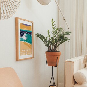 May include: A framed poster with a colorful illustration of a wave breaking at "The Wedge" in Newport Beach, California. The poster is hanging on a white wall next to a potted plant and a gold mirror.