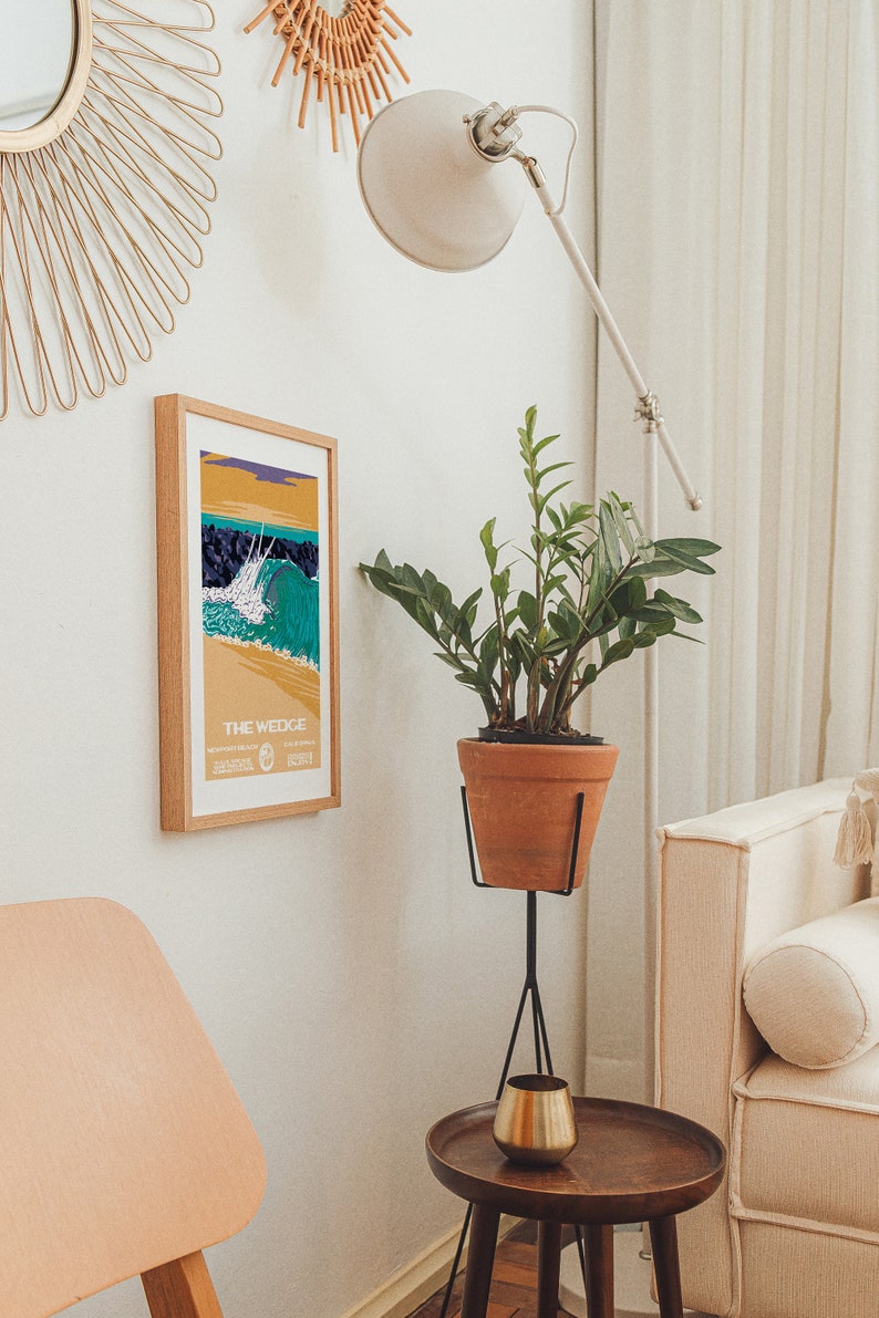 May include: A framed poster with a colorful illustration of a wave breaking at "The Wedge" in Newport Beach, California. The poster is hanging on a white wall next to a potted plant and a gold mirror.