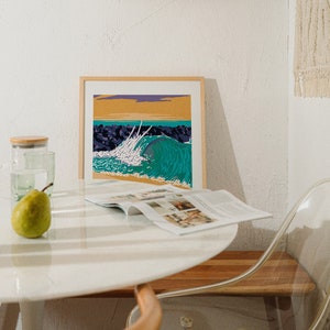 May include: A framed print of a colorful ocean wave crashing against a rocky shore. The print is hanging on a white wall above a white table with a magazine and a green pear on it.