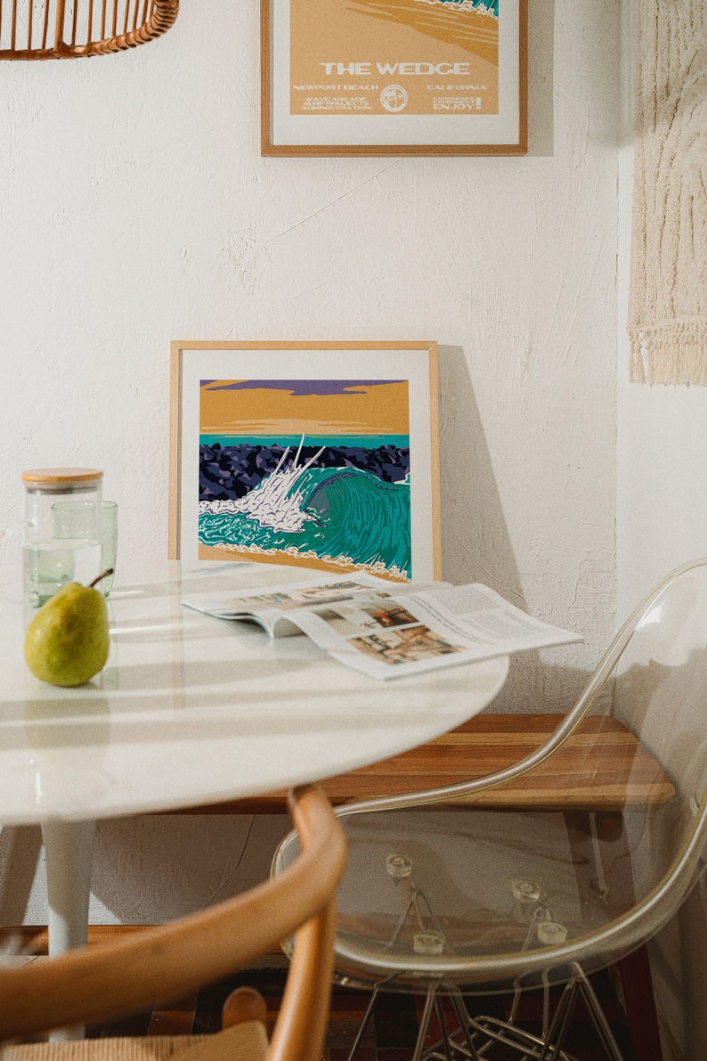 May include: A framed print of a colorful ocean wave crashing against a rocky shore. The print is hanging on a white wall above a white table with a magazine and a green pear on it.