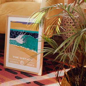 May include: A framed poster with a vintage style illustration of a large wave breaking at "The Wedge" in Newport Beach, California. The poster is in a wooden frame and is sitting on a red and black rug.