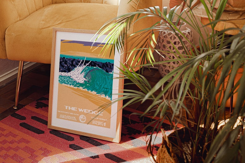 May include: A framed poster with a vintage style illustration of a large wave breaking at "The Wedge" in Newport Beach, California. The poster is in a wooden frame and is sitting on a red and black rug.