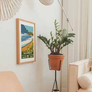 May include: A white lamp with a white shade is positioned in a corner of a room. A potted plant with green leaves sits on a black stand. A framed poster with the text "DEVEREUX, LA VISTA, CALIFORNIA" is on the wall. A wooden side table with a gold cup sits next to a white couch.