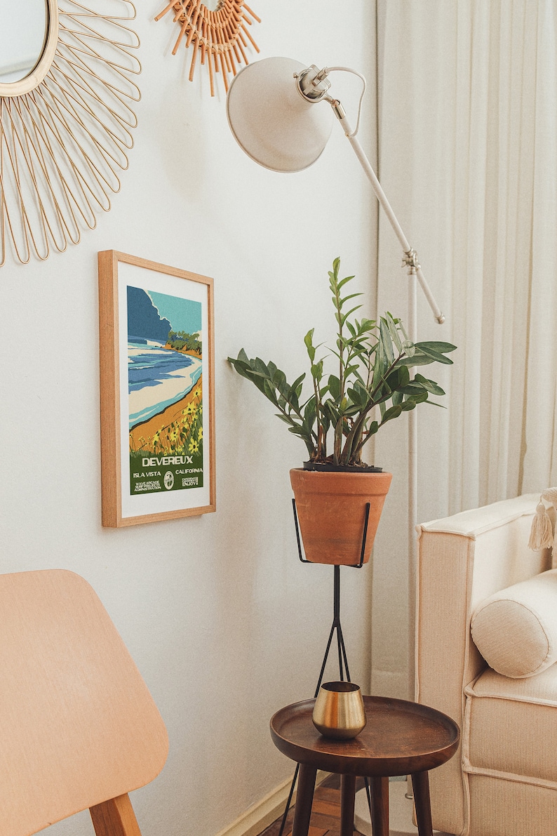 May include: A white lamp with a white shade is positioned in a corner of a room. A potted plant with green leaves sits on a black stand. A framed poster with the text "DEVEREUX, LA VISTA, CALIFORNIA" is on the wall. A wooden side table with a gold cup sits next to a white couch.