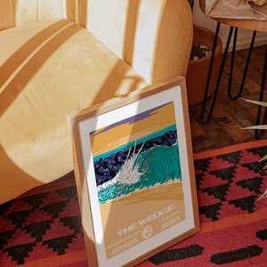 May include: A framed print of a vintage style illustration of a large wave breaking at The Wedge in Newport Beach, California. The print is in a wooden frame and is sitting on a red and black rug.