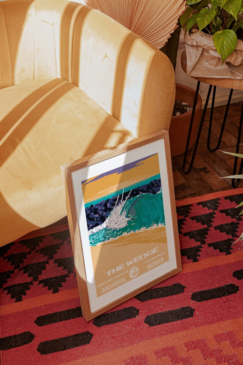 May include: A framed print of a vintage style illustration of a large wave breaking at The Wedge in Newport Beach, California. The print is in a wooden frame and is sitting on a red and black rug.