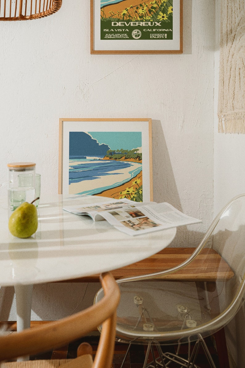 May include: A white table with a magazine, a green pear, and a glass of water. A framed print of a beach scene hangs on the wall above the table. A clear plastic chair sits next to the table.