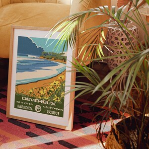 May include: A framed vintage travel poster with a blue, green, and yellow color scheme. The poster features a beach scene with a blue ocean, a sandy beach, and a green hillside. The text on the poster reads "Devereux, Isla Vista, California, Enjoy!"
