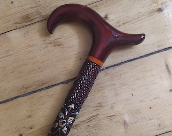 May include: A dark brown wooden cane with a curved handle. The handle and upper shaft are a glossy, rich brown. The lower shaft features intricate carved floral and geometric patterns, with a contrasting orange band.