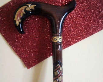 May include: A wooden walking cane with a curved handle. The handle is dark brown with gold and black decorative accents. The shaft is also dark brown with carved gold and copper-colored designs. The cane is a stylish mobility aid.