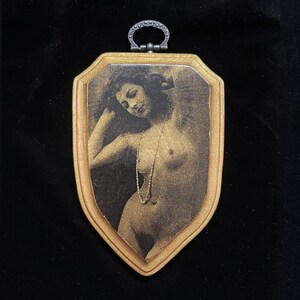 May include: A gold-toned framed picture with a black and white image of a woman with long hair wearing a pearl necklace. The frame has a decorative metal loop at the top.