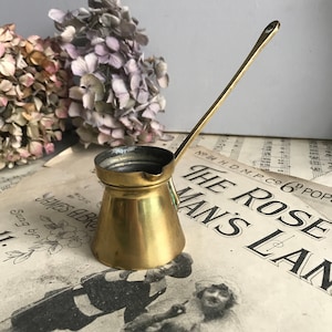 May include: A small, vintage brass coffee pot with a long handle. The pot has a rounded base and a narrow spout. The background includes sheet music and dried flowers, suggesting a rustic or antique aesthetic.