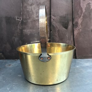 May include: A vintage brass bucket with a curved handle. The bucket has a rounded shape and a shiny, golden finish. The handle is made of a darker metal and is attached to the bucket with copper rivets. The bucket is likely used for carrying water or other items.