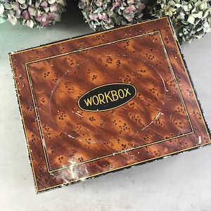 May include: A vintage rectangular workbox with a faux wood grain pattern in shades of brown. The box has a black border with gold trim and the word "WORKBOX" in a black oval with gold lettering.