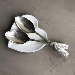 May include: Two silver spoons rest on a white, heart-shaped dish. The spoons have ornate detailing on the handles. The dish is set against a gray background.