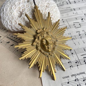 May include: A vintage brass sunburst pendant featuring a detailed face at the center. The pendant has radiating rays and a small crown. The background includes sheet music and a textured white object.