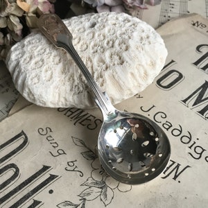 May include: A silver tea strainer spoon with a perforated bowl and a decorative handle. The spoon is resting on a vintage sheet music background. The handle has an embossed design. The spoon is likely made of sterling silver.