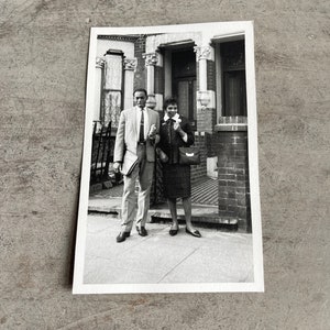 May include: Black and white photograph of a formally dressed couple posing in front of a brick building. The man wears a suit and tie, holding a notebook. The woman wears a skirt suit and carries a purse. The image is printed on a white rectangular card.