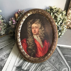 May include: A vintage oval-shaped tray with a gold-painted frame and a portrait of a man in a red and white outfit. The man has long, curly, blonde hair and is wearing a white lace collar.