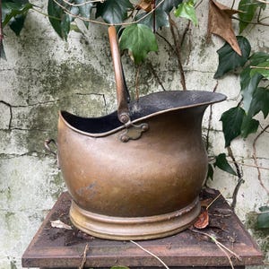 Puede incluir: Un antiguo balde de carbón de cobre con un asa de madera y una tapa con bisagras. El balde tiene un cuerpo redondeado y una abertura ancha, con un interior oscuro. Se encuentra sobre una superficie metálica contra una pared desgastada con hiedra.