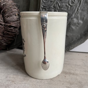 May include: A white ceramic mug with a silver spoon hanging on the rim. The spoon has a decorative design on the handle. The mug has some text on the side. The background is blurred.