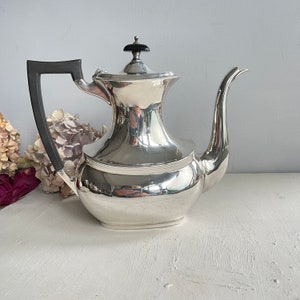 May include: A silver coffee pot with a black handle and lid finial. The pot has a curved spout and a rounded body. The handle is a dark, rectangular shape. The pot is on a white surface. The pot's design is Art Deco inspired.