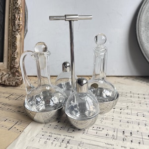 May include: A silver condiment set featuring four clear glass bottles. Two bottles have handles, and all have silver stoppers. The set is supported by a silver metal stand. The bottles rest on vintage sheet music.