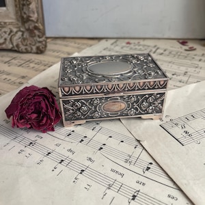 May include: A silver trinket box with an ornate floral design and a mirrored interior. The box is sitting on a sheet of music.
