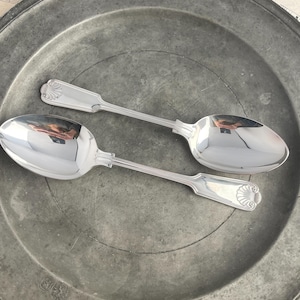 Puede incluir: Dos cucharas de plata con mangos adornados y un acabado pulido. Las cucharas están sobre un plato de peltre.